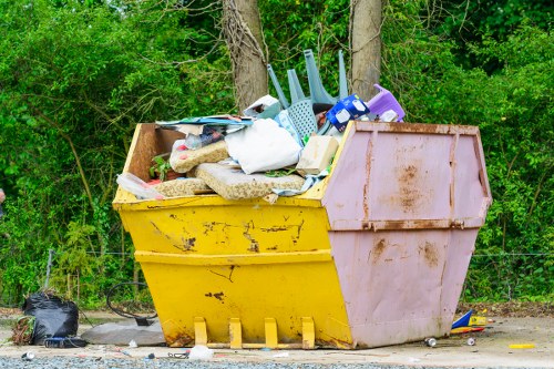 A large open-top skip bin placed outdoors on a gravel surface, filled with various waste items including plastic and metal objects, some household items like brushes and containers, as well as textiles and cardboard packaging. The bin is yellow on the front with a faded pink back section, showing signs of rust and wear. It is positioned near a background of green foliage and trees, with a black garbage bag and scattered debris on the ground nearby. The scene appears to be part of a waste disposal or rubbish removal site in Shepherds Bush, with the environment suggesting a suburban or urban setting dedicated to waste management services provided by Commercial Waste Shepherds Bush.
