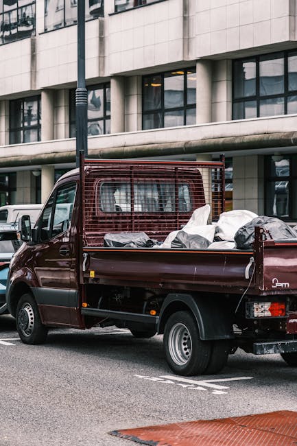 A small burgundy flatbed truck parked on a city street in front of a modern building with large glass windows and concrete exterior. The truck's bed is loaded with various mixed waste materials, inclu