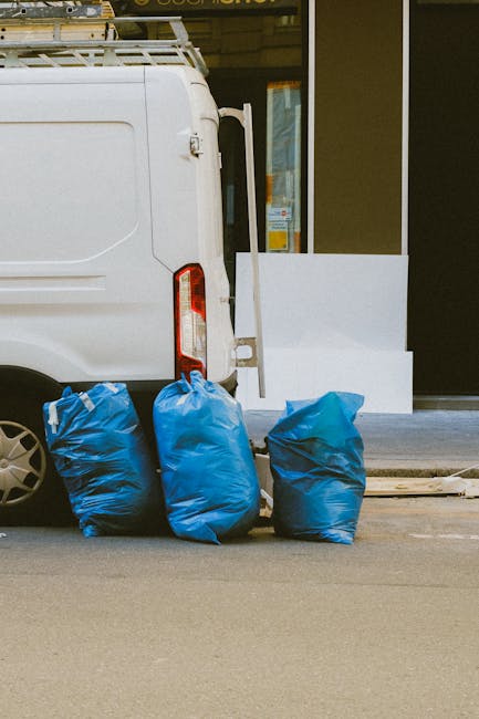 A small burgundy flatbed truck parked on a city street in front of a modern building with large glass windows and concrete exterior. The truck's bed is loaded with various mixed waste materials, inclu